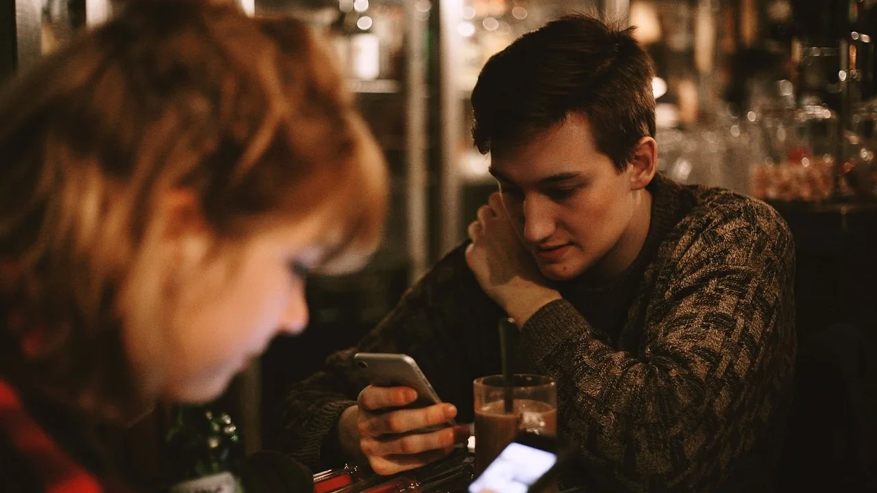 Two people at a dimly lit bar, each looking at their smartphone, with drinks on the table.
