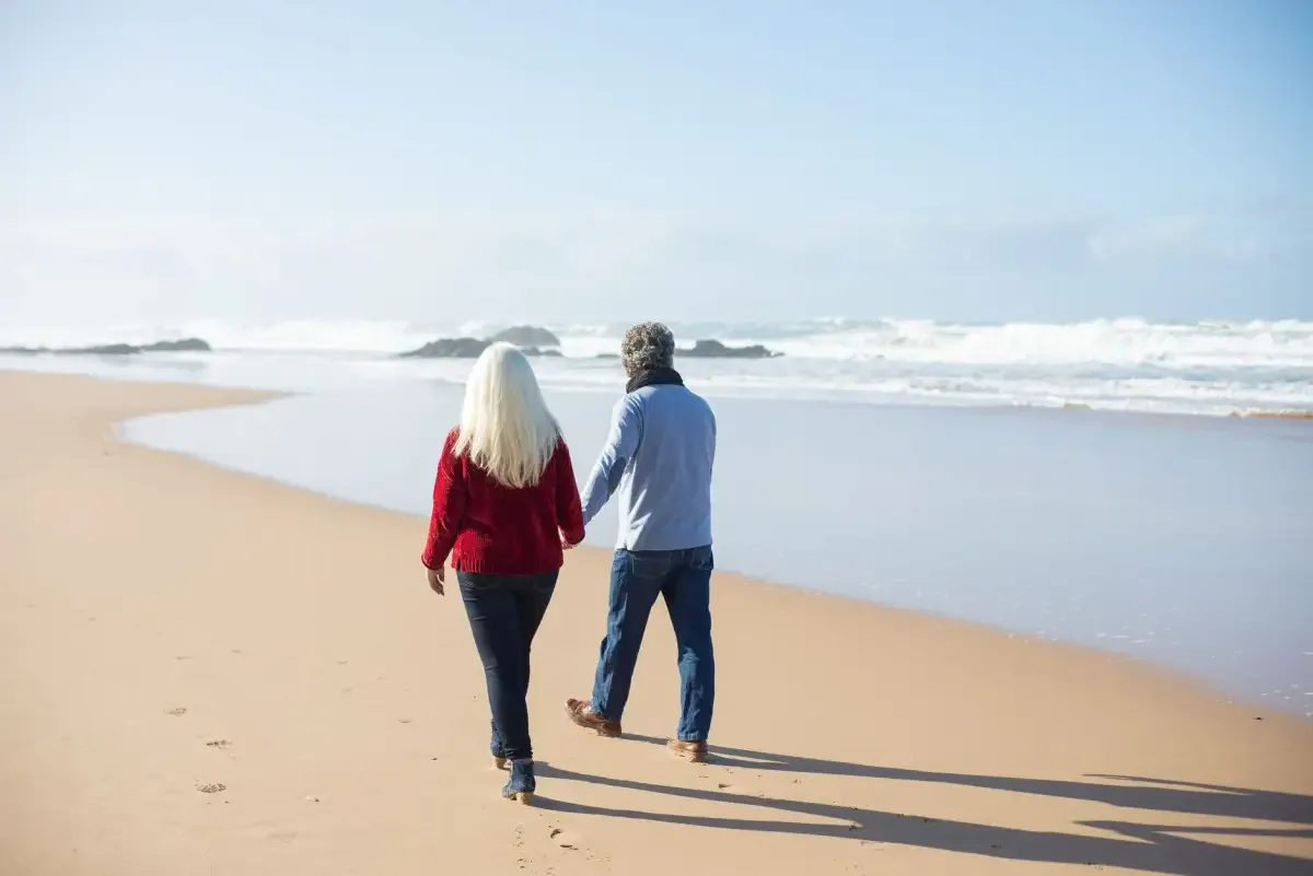 An older couple walking hand in hand along a sunny beach, seen from behind